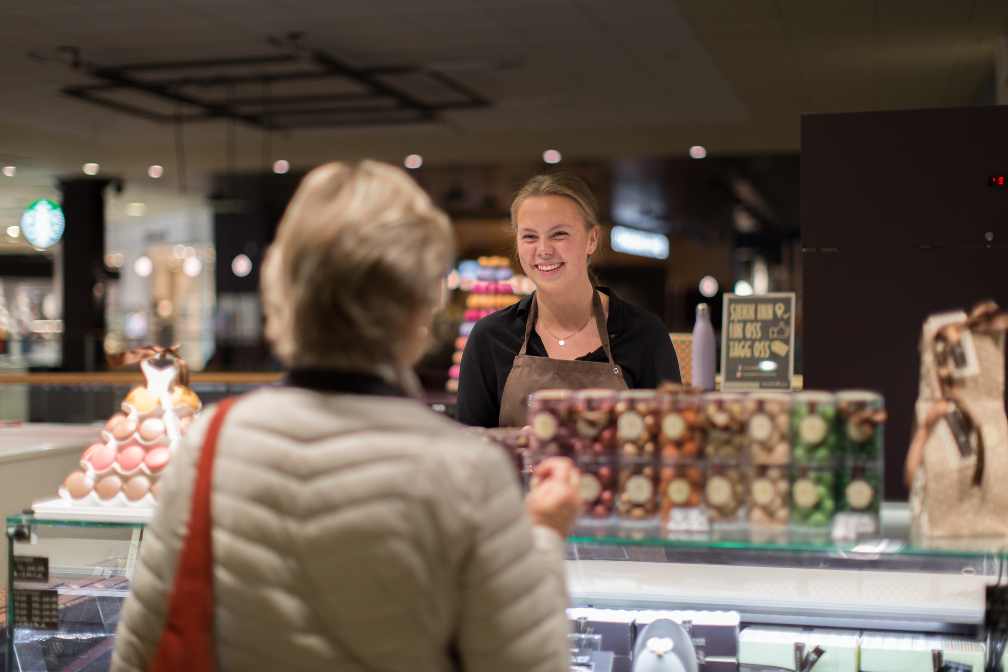 Bilde av en eldre dame som står ved en handledisk og en smilende ekspeditør hjelper henne. 